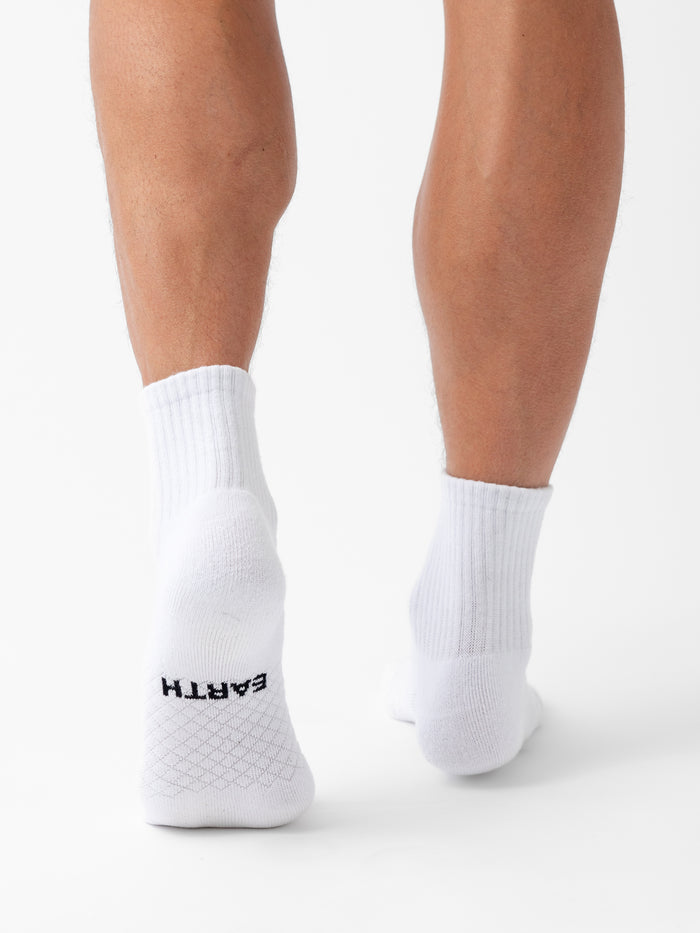 A person wearing Cozy Earth Essential Quarter Socks stands on a white background, back of legs visible. The word "EARTH" is printed in black near the heel on the sole of the left sock. |Color:White