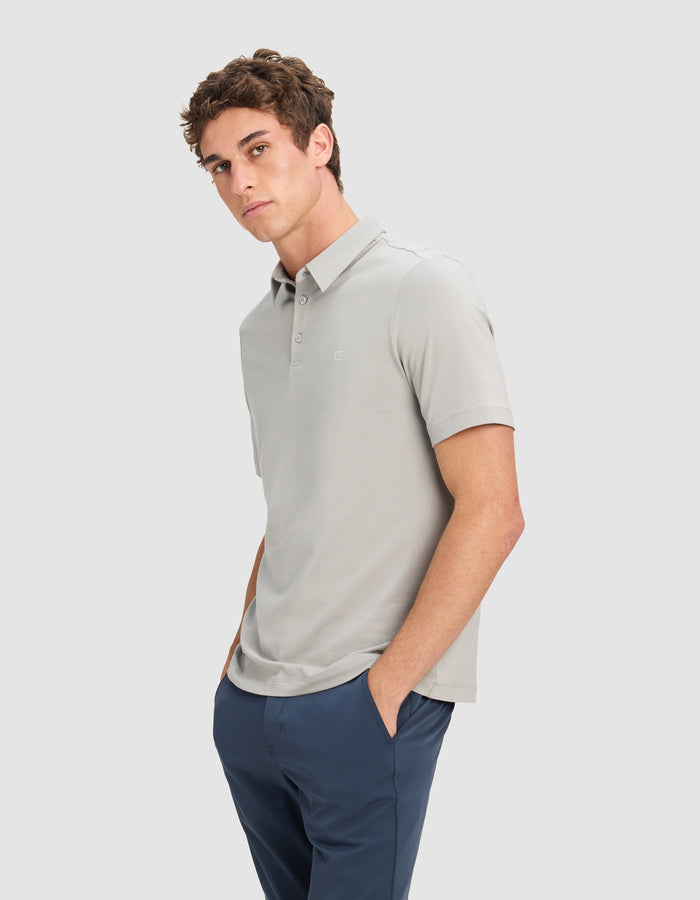 A man with short, curly brown hair wears the Cozy Earth Men's Everyday Polo in light gray and navy pants. He stands against a plain light background, hands in his pockets, gazing slightly to the side.|Color:Stone