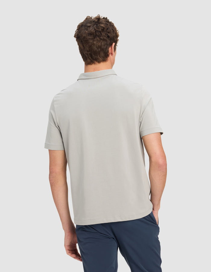 A man with short curly hair is shown from the back wearing the Cozy Earth Men's Everyday Polo in light gray and dark blue pants, standing against a plain light background.|Color:Stone