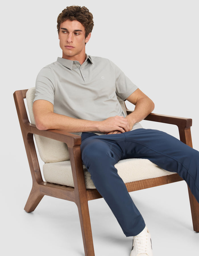 A man wearing the Cozy Earth Men's Everyday Polo sits on a cushioned wooden chair, paired with navy pants and white sneakers, looking off to the side against a plain background.|Color:Stone