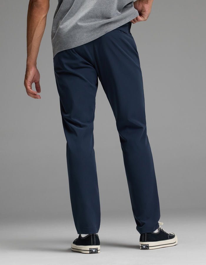 A man stands facing away against a plain background, wearing the Cozy Earth Men’s Everywhere Pant in navy blue, paired with a grey shirt and black sneakers with white soles, one hand resting on his hip.|Color:Eclipse