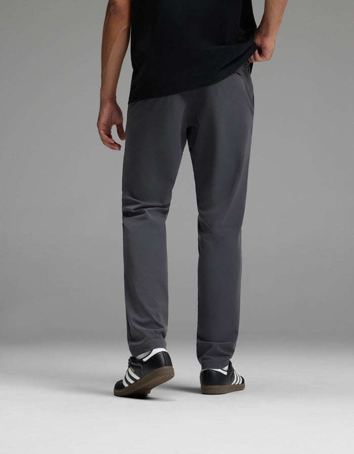 A person stands facing away, wearing a black shirt, Cozy Earth Men’s Everywhere Pant in gray, and black-and-white Adidas sneakers, against a plain gray background.|Color:Graphite