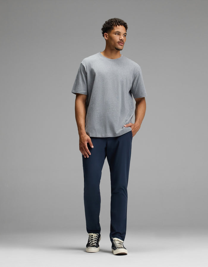 A man stands against a gray background, wearing the Cozy Earth Men’s Everywhere Pant in dark blue, paired with a light gray t-shirt and black-and-white sneakers. One hand is in his pocket as he looks slightly to the side with a neutral expression.|Color:Eclipse