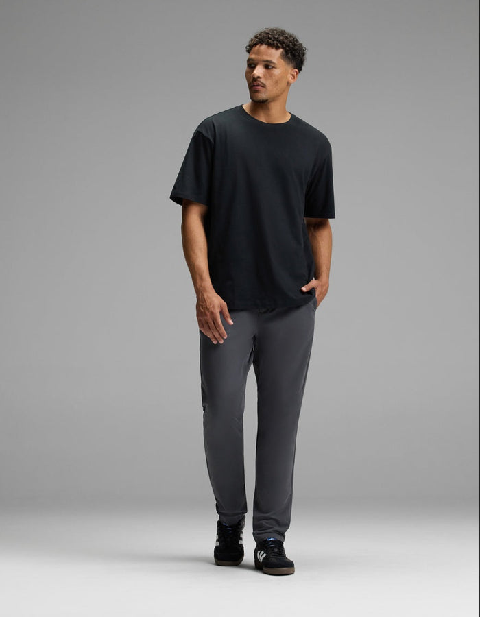 A man stands against a plain gray background, wearing Cozy Earth’s Men’s Everywhere Pant in gray, paired with a black T-shirt and black shoes. He has short curly hair and is looking slightly to his right, with one hand in his pocket.|Color:Graphite