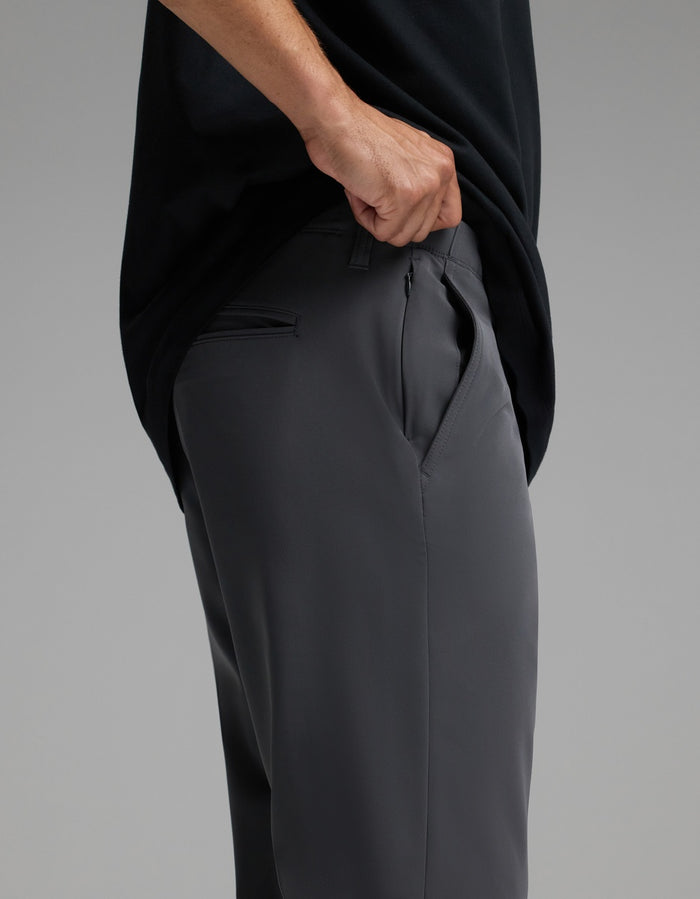 A person wearing Cozy Earth’s Men’s Everywhere Pant in dark gray and a black shirt lifts their shirt to reveal the side zipper pocket on the pants. The background is plain and gray.|Color:Graphite