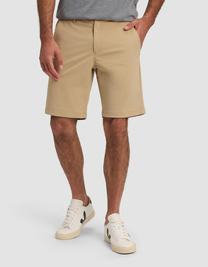 A man wearing Cozy Earth Men's Everywhere Short in beige, a gray t-shirt, and white sneakers with black accents stands with one hand in his pocket against a plain background.|Color:Sandalwood