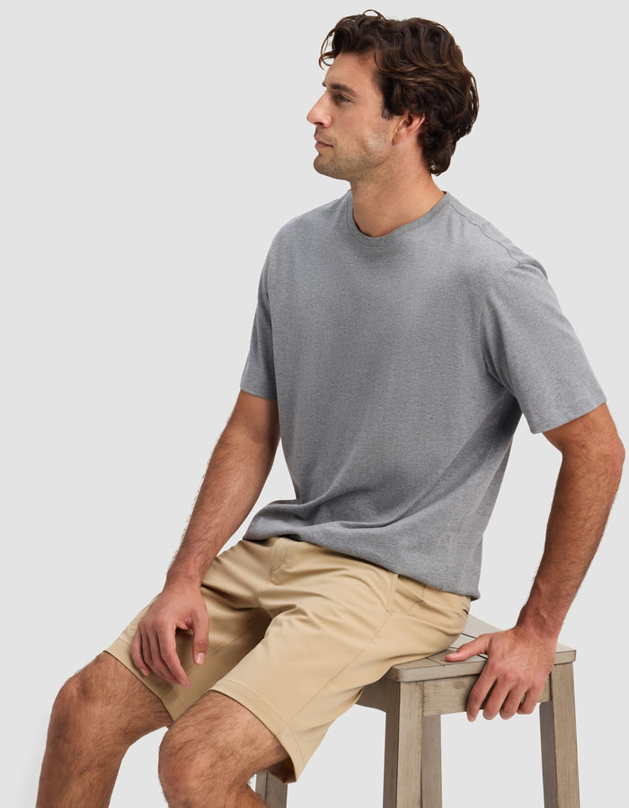 A man with wavy brown hair, wearing a gray t-shirt and Cozy Earth Men's Everywhere Short in beige, sits on a wooden stool against a plain light background, looking to his left.|Color:Sandalwood