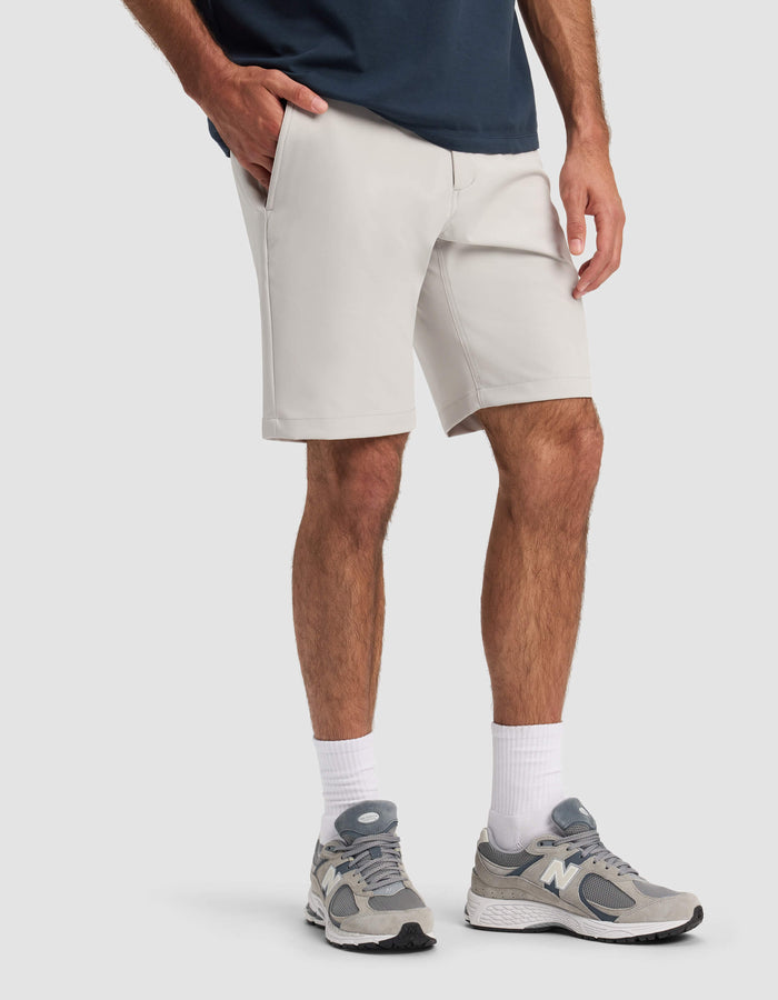 A person wearing Cozy Earth Men's Everywhere Short in light gray, a navy shirt, white crew socks, and gray New Balance sneakers stands with one hand in their pocket against a plain light background.|Color:Dew