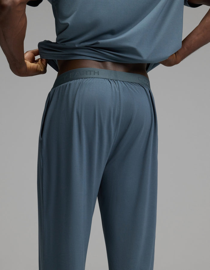 A person is wearing the Cozy Earth Men’s First Layer Pajama Pant, paired with a blue-gray shirt. They are lifting their shirt slightly to reveal the waistband of the pajama pants. The background is plain gray.|Color:Dawn