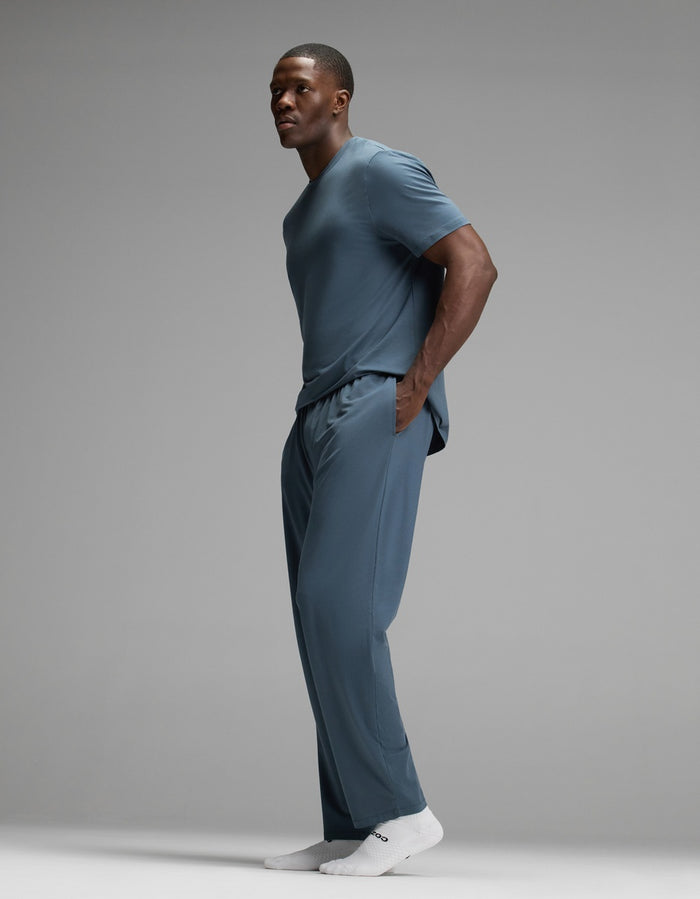 A man stands sideways against a plain gray background, wearing the Cozy Earth Men’s First Layer Pajama Pant in blue as part of a matching set with a blue t-shirt and white socks. He has his hands in his pockets.|Color:Dawn