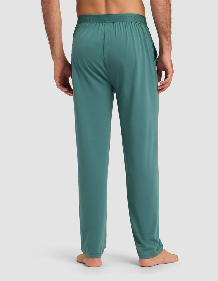 A man stands barefoot, back facing the camera, in Cozy Earth Men's First Layer Pajama Pant. The relaxed-fit teal pants feature side pockets and a subtly branded waistband, set against a plain light gray background.|Color:Sea Green