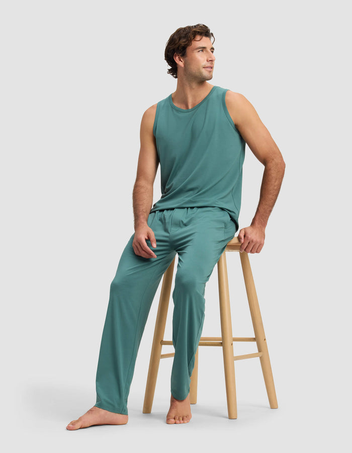 A man in Cozy Earth's Men's First Layer Pajama Pant, paired with a teal sleeveless top, sits barefoot on a wooden stool, smiling and looking left against a plain light background.|Color:Sea Green
