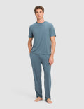 A person stands barefoot against a plain background, wearing Cozy Earth's Men's First Layer Pajama Pant in blue with a matching top. Their left hand is in a pocket, and they face forward with a neutral expression.|Color:Dawn