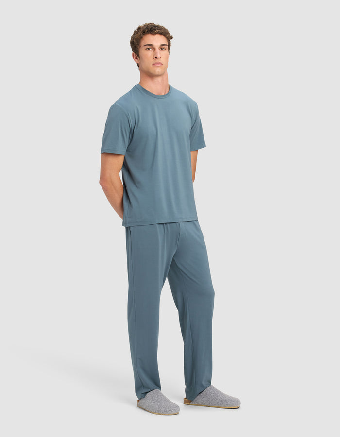 A man with short curly hair stands against a light background wearing the Cozy Earth Men's First Layer Pajama Short Sleeve Tee & Pant Set in blue and gray slippers, with his hands behind his back.|Color:Dawn