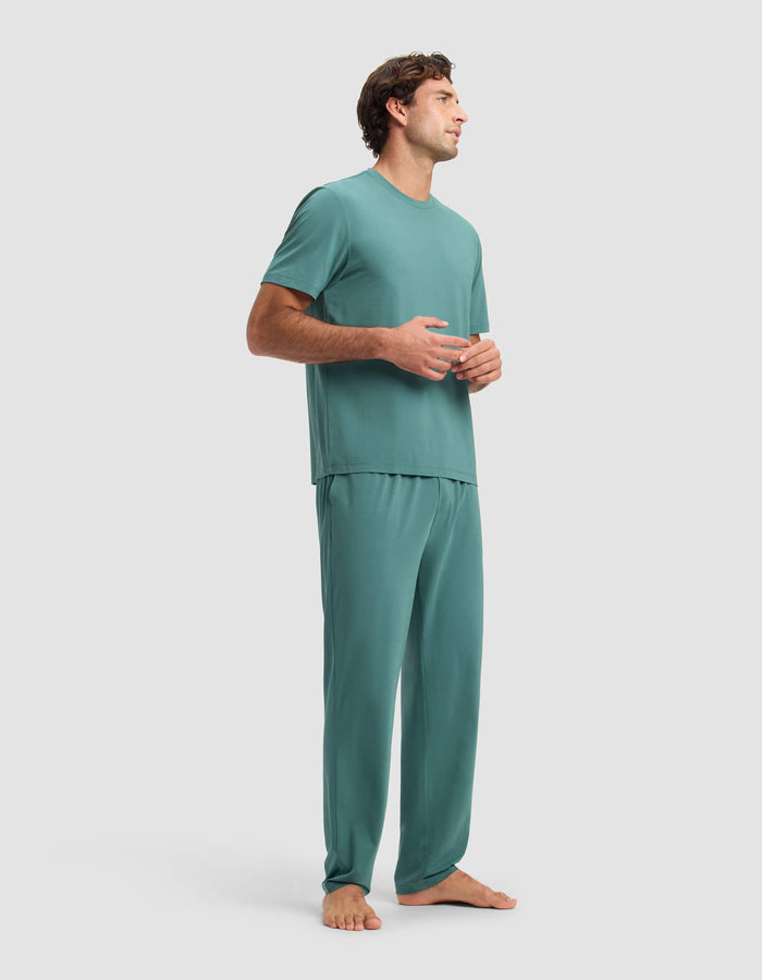 A man stands barefoot on a light background, wearing the Cozy Earth Men's First Layer Pajama Short Sleeve Tee & Pant Set in teal. He looks to the side with his hands loosely clasped.|Color:Sea Green