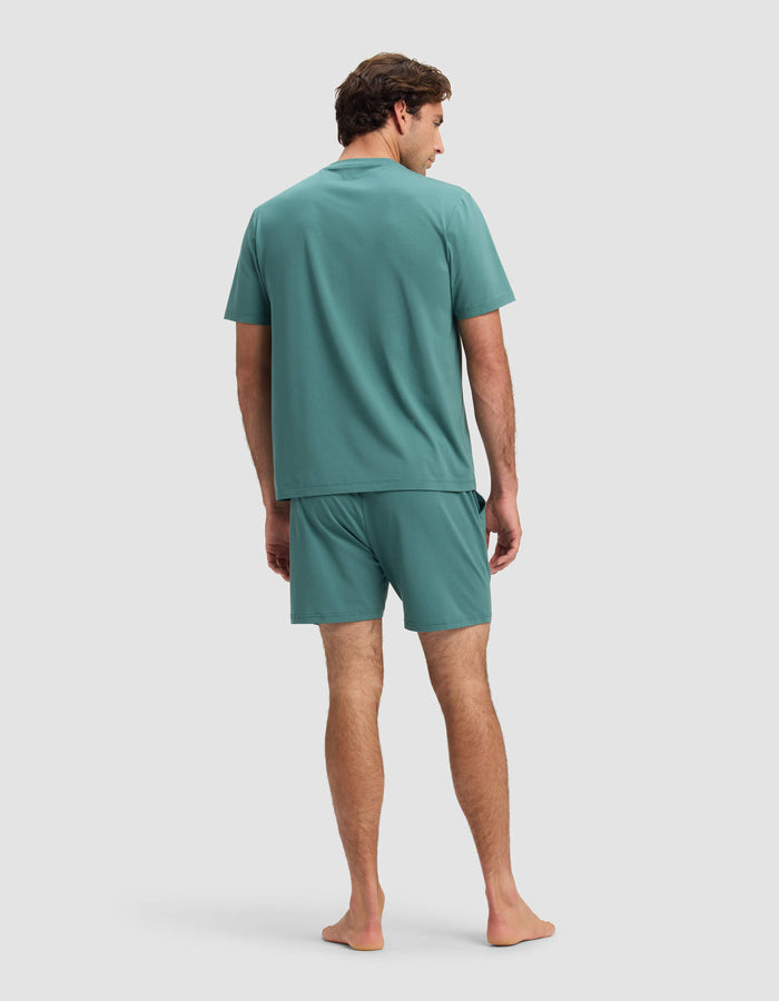 A man stands barefoot facing away, wearing the Cozy Earth Men's First Layer Pajama Short Sleeve Tee & Short Set in teal, against a plain light gray background.|Color:Sea Green