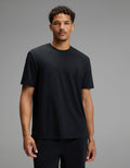 A man with short curly hair wears the Cozy Earth Men’s First Layer Pajama Short Sleeve Tee in Jet Black along with black pants, standing against a plain gray background. He faces forward with a neutral expression.|Color:Jet Black