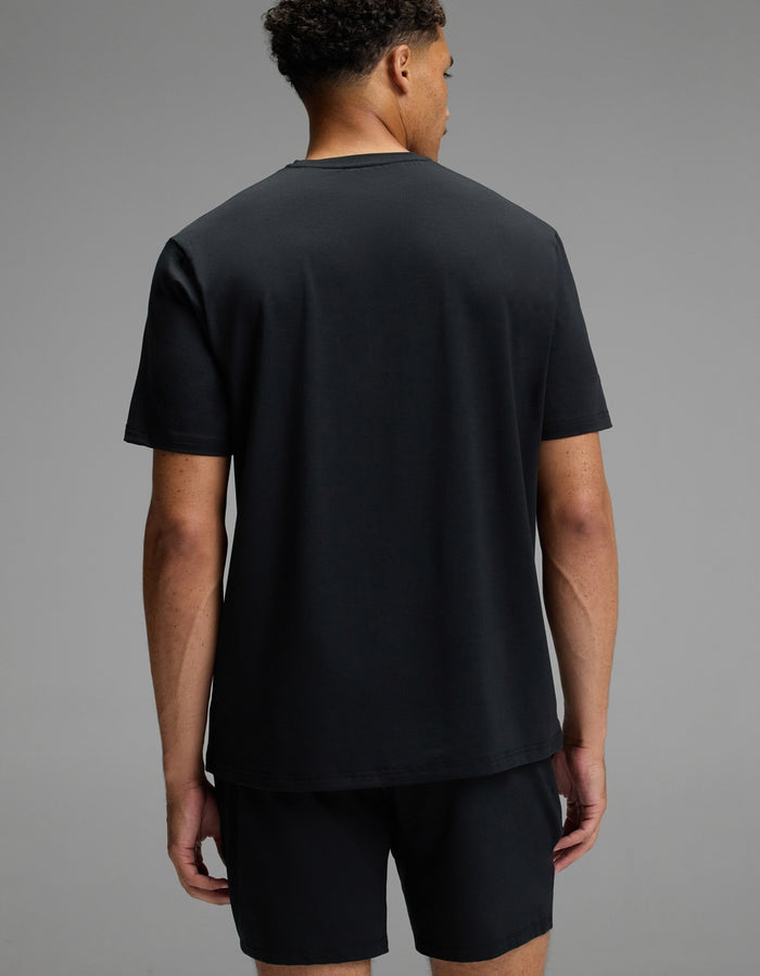 A man stands facing away from the camera, wearing the Cozy Earth Men’s First Layer Pajama Short Sleeve Tee in black and matching black shorts against a plain gray background.|Color:Jet Black