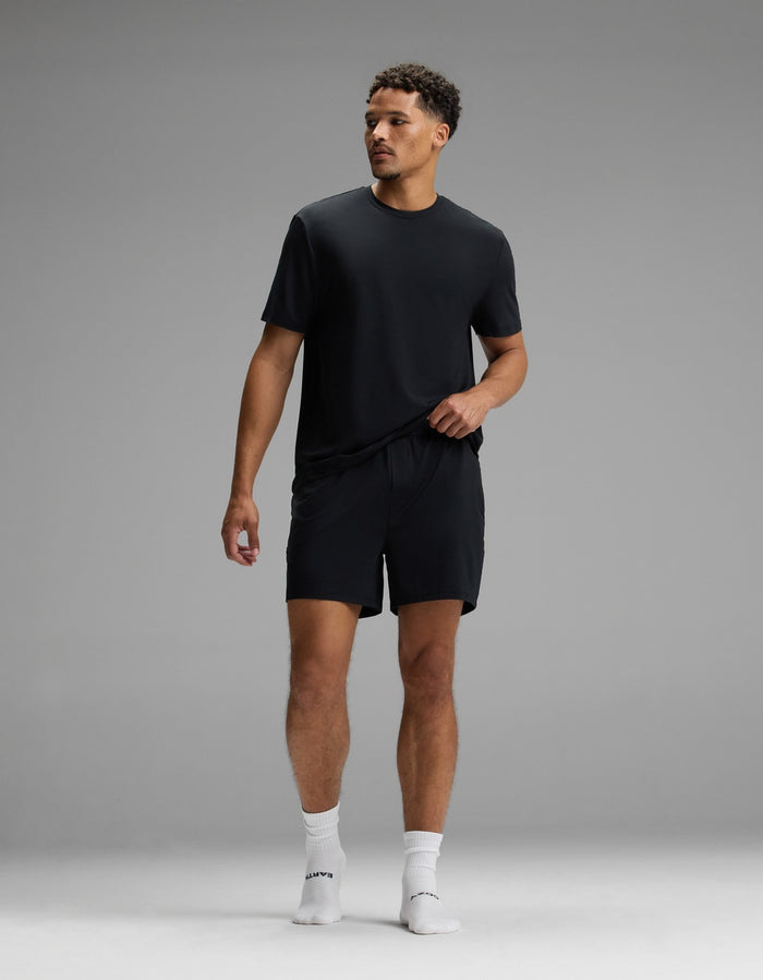 A man stands in a studio against a plain gray background, wearing the Cozy Earth Men’s First Layer Pajama Short Sleeve Tee in black, paired with black shorts and white socks. He looks to his right with one hand by his side and the other touching his shorts.|Color:Jet Black