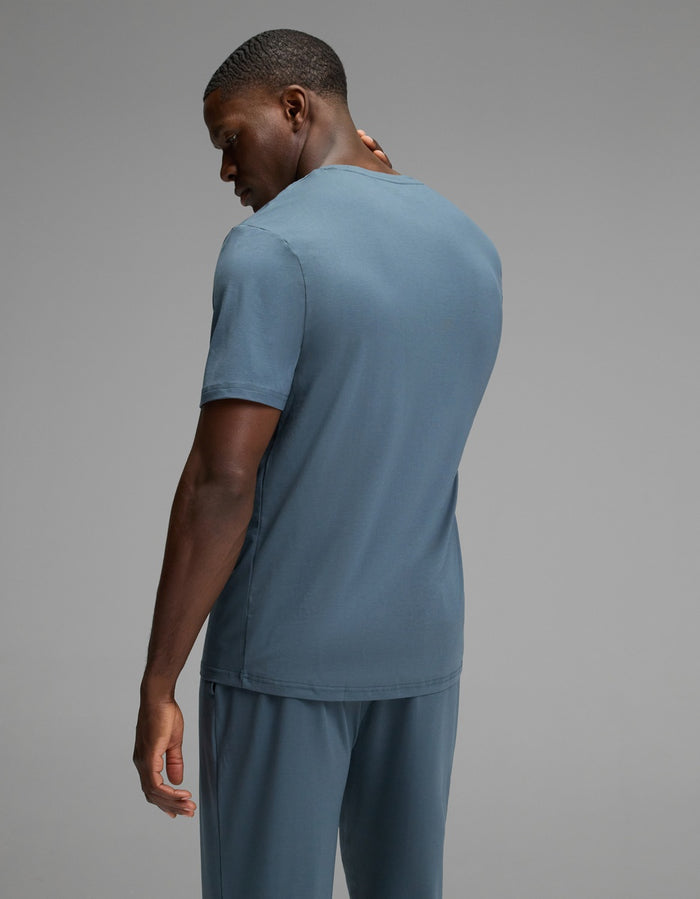 A man models the Cozy Earth Men’s First Layer Pajama Short Sleeve Tee in blue with matching pants, standing against a plain gray background, his head turned slightly to the side.|Color:Dawn