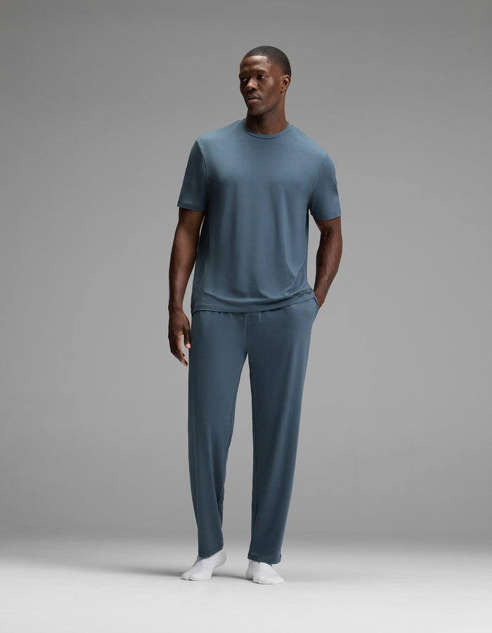 A man stands against a gray background, wearing the Cozy Earth Men’s First Layer Pajama Short Sleeve Tee in blue-gray, paired with matching pants and white socks. His right hand rests at his side while his left hand is tucked into his pocket.|Color:Dawn