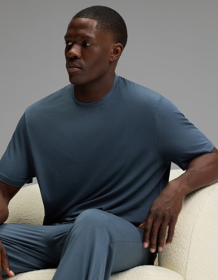 A man wearing the Cozy Earth Men’s First Layer Pajama Short Sleeve Tee sits on a light-colored chair, looking slightly to his left with a neutral expression. The background is plain and gray.|Color:Dawn