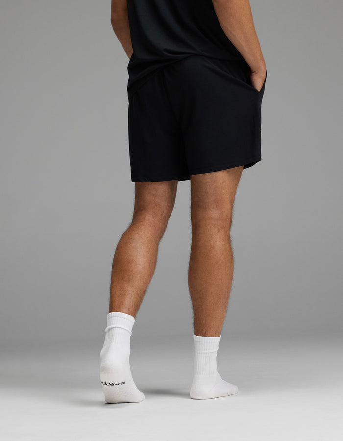 A person stands with hands in pockets, wearing the Cozy Earth Men’s First Layer Pajama Short in black, paired with a black shirt and white socks. The image displays their back and legs against a plain gray background.|Color:Jet Black
