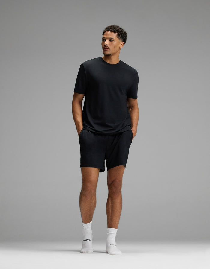 A man stands against a plain gray background, wearing Cozy Earth’s Men’s First Layer Pajama Short, paired with a black t-shirt, white crew socks, and white sneakers. His hands are in his pockets as he looks off to the side.|Color:Jet Black