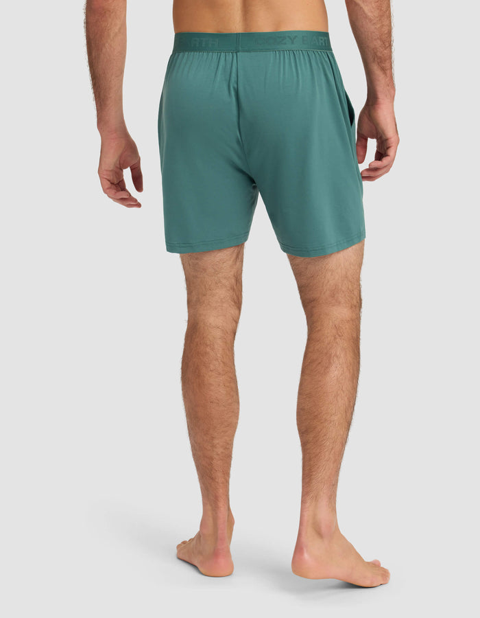 A man stands barefoot on a white background, seen from the back, wearing Cozy Earth's Men's First Layer Pajama Short in teal, featuring an elastic waistband and side pockets.|Color:Sea Green