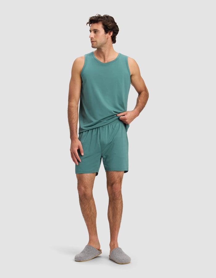 A man with short dark hair stands against a plain background wearing Cozy Earth's Men's First Layer Pajama Short set in teal and gray slippers, looking slightly to his left.|Color:Sea Green