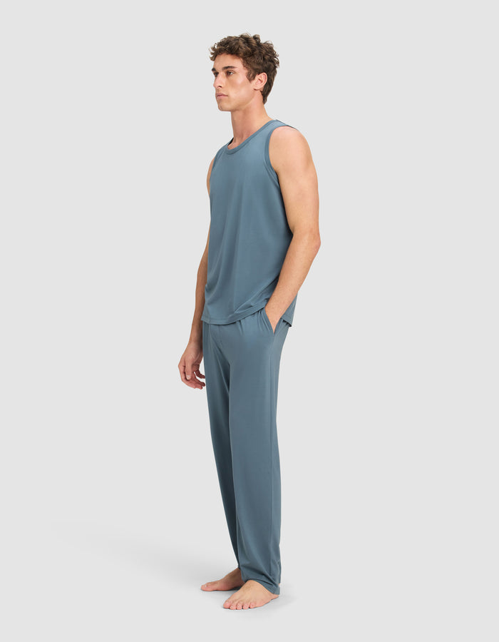 A man stands barefoot against a light gray background, wearing Cozy Earth's Men's First Layer Pajama Tank & Pant Set in blue-gray, with his hands in his pockets.|Color:Dawn