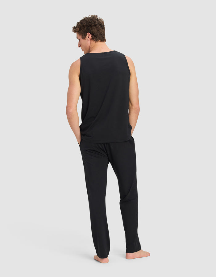 A man stands barefoot, wearing the Cozy Earth Men's First Layer Pajama Tank & Pant Set in black, with his hands in his pockets, facing away from the camera against a plain light gray background.|Color:Jet Black