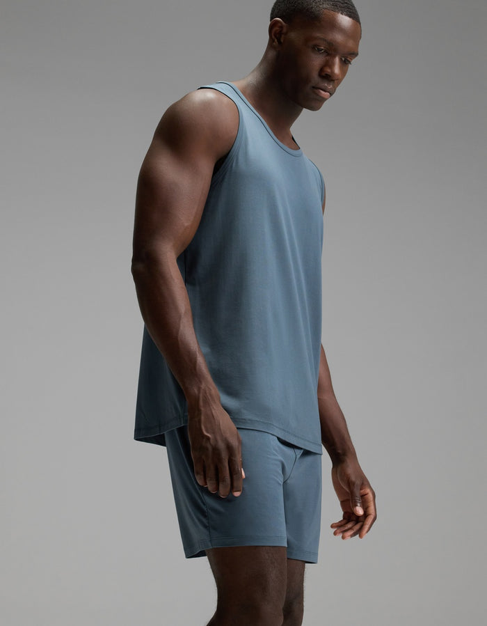 A man stands in a studio wearing the Cozy Earth Men’s First Layer Pajama Tank and matching shorts in blue, looking downward with a neutral expression against a plain gray background.|Color:Dawn