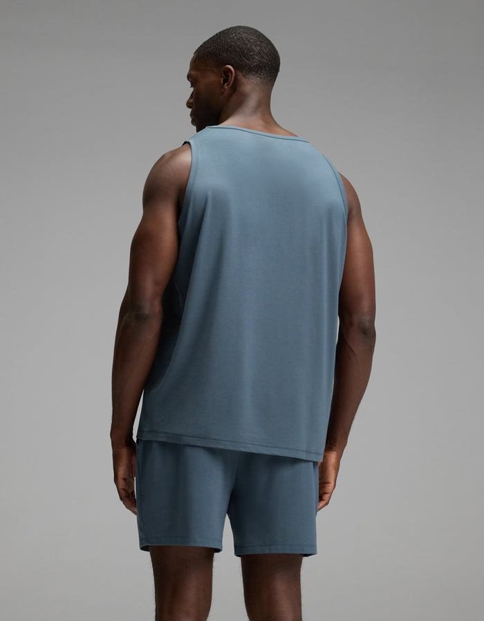 A man stands facing away from the camera, wearing the Cozy Earth Men’s First Layer Pajama Tank in blue, paired with matching blue shorts, against a plain gray background.|Color:Dawn