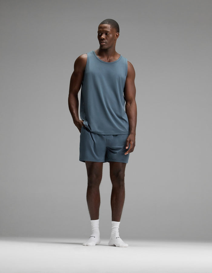 A man wearing the Cozy Earth Men’s First Layer Pajama Tank in blue, with matching shorts and white socks, stands against a plain gray background, looking to his left with one hand in his pocket.|Color:Dawn