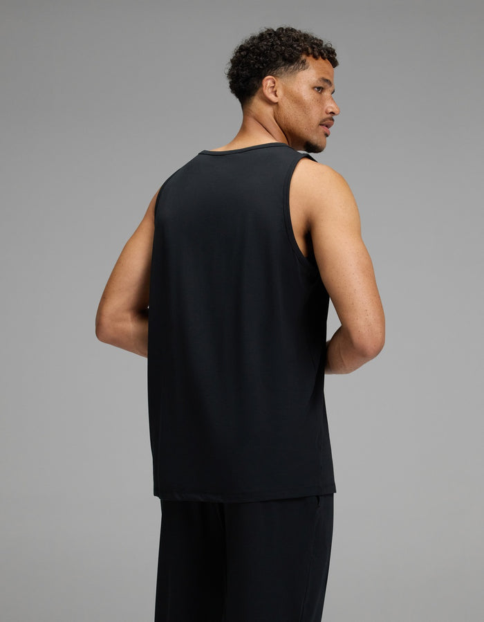A man with short curly hair is wearing the Cozy Earth Men’s First Layer Pajama Tank in black and matching black pants, facing away from the camera against a plain grey background.|Color:Jet Black