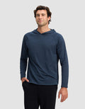 A man wearing Cozy Earth's Men's Lightweight Performance Hoodie in dark blue and black pants stands against a plain light gray background, one hand in his pocket and a slight smile.|Color:Heather Eclipse