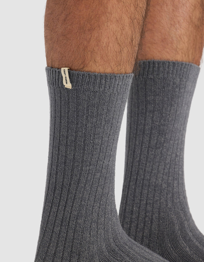 Close-up of a person's lower legs wearing The Plush Lounge Calf Sock in dark gray from Cozy Earth, featuring ribbed texture and a small beige tag near the top. Plain white background.|Color:Charcoal/Coal/Stone