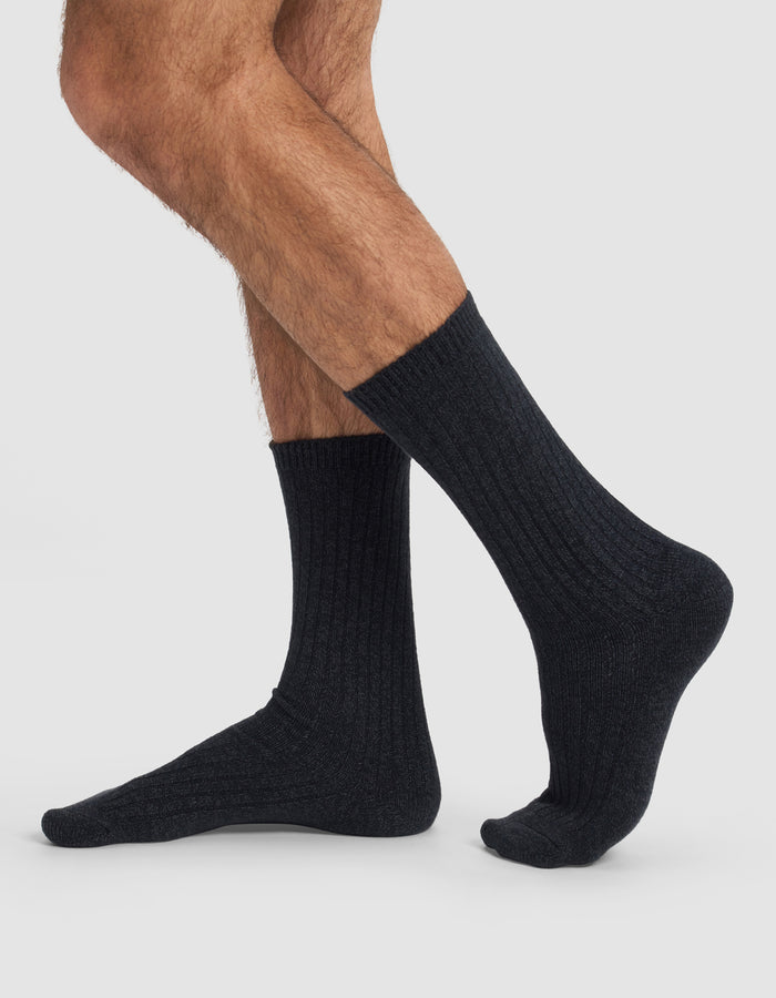 A person with crossed legs wears Cozy Earth's The Plush Lounge Calf Sock 3-Pack, dark ribbed crew socks, photographed from the side against a plain light background.|Color:Charcoal/Coal/Stone