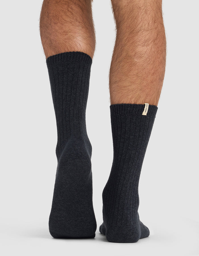 A person in Cozy Earth’s The Plush Lounge Calf Sock 3-Pack stands on a light surface, seen from behind at ankle level. The left heel is slightly raised, and a small tag is visible on the right sock.|Color:Charcoal/Coal/Stone