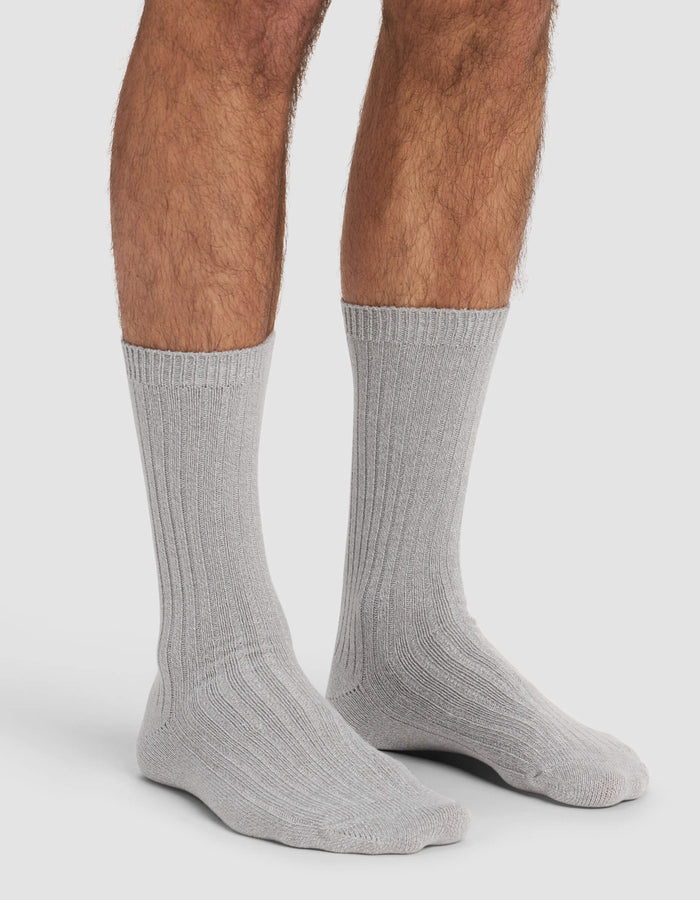 A person wears Cozy Earth's Men's Plush Lounge Calf Socks in light gray, standing on a white background with only their lower legs and feet visible.|Color:Stone