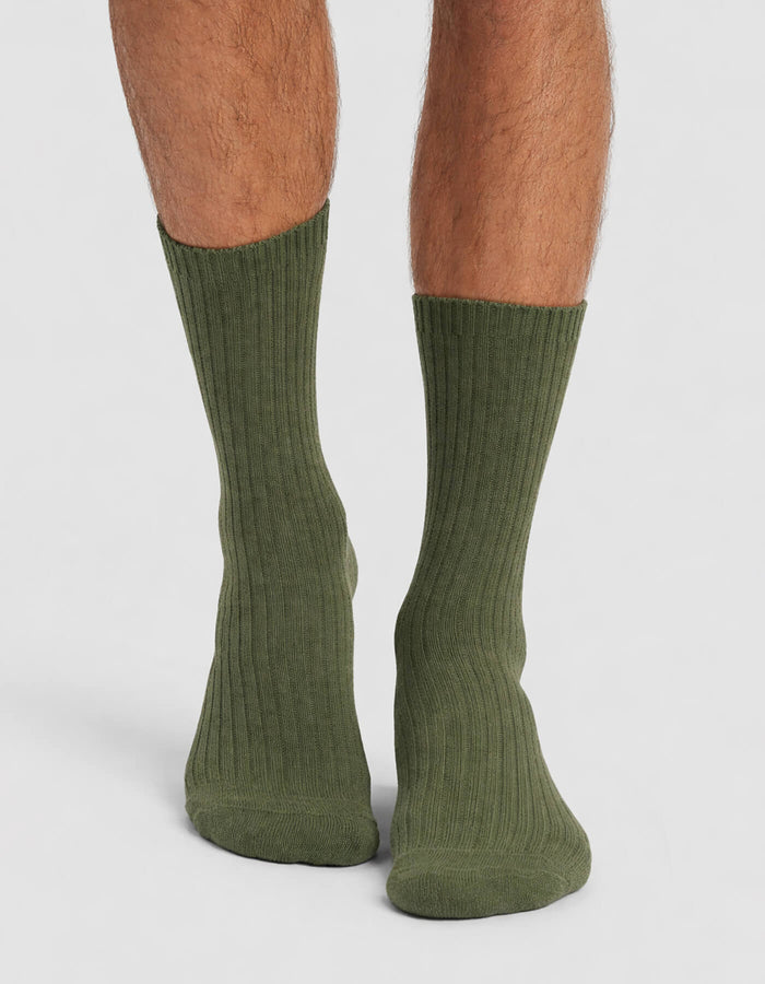 A person wears Cozy Earth's Men's Plush Lounge Calf Sock in ribbed olive green, standing on a light gray surface with the socks shown from back and side angles.|Color:Olive