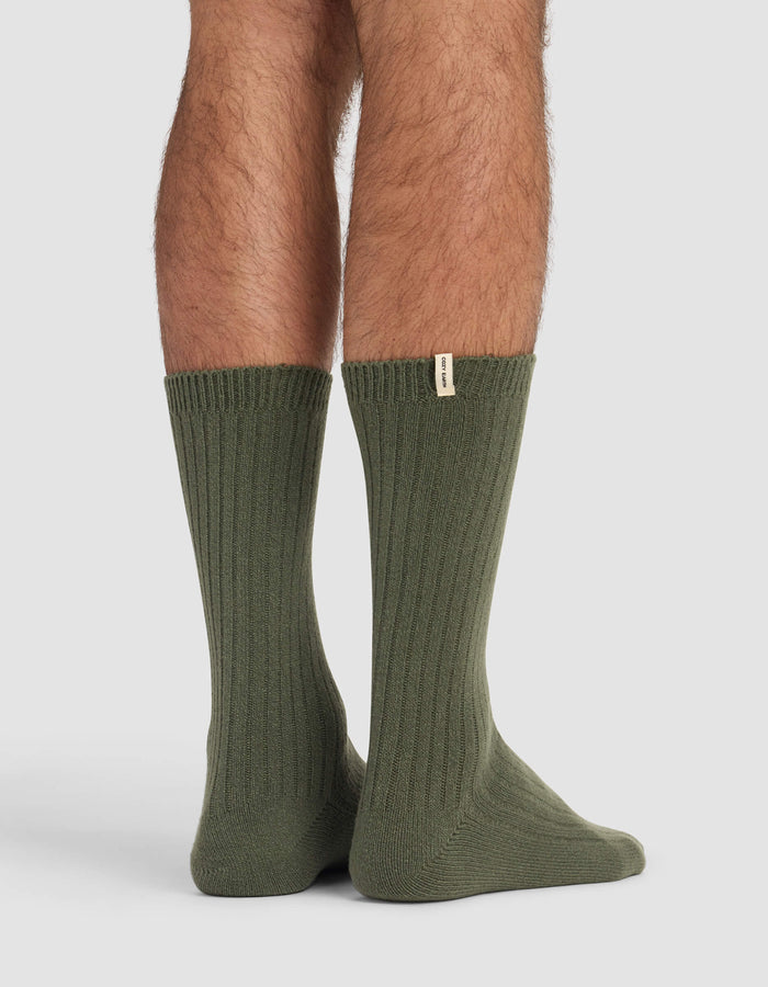 A person stands on a light surface wearing Cozy Earth's Men's Plush Lounge Calf Sock in olive green, featuring ribbing and a small beige tag near the top. Only their lower legs and feet are visible.|Color:Olive