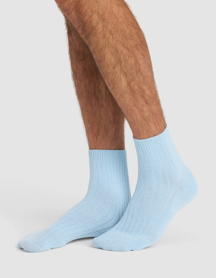 A person with crossed legs is wearing Cozy Earth’s Plush Lounge Quarter Sock 3-Pack in light blue, featuring a ribbed crew design against a plain white background.|Color:Pacific/Harbor/Lake