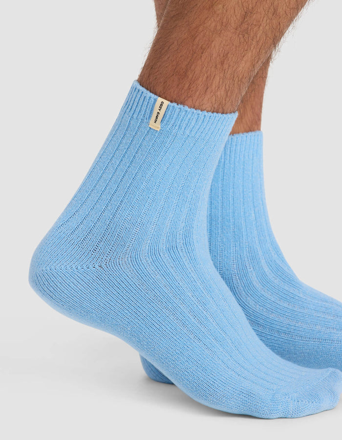 Close-up of a person wearing Cozy Earth Men's Plush Lounge Quarter Socks in light blue with ribbed texture and a small beige tag near the cuff. The background is plain white. Part of a 3-pack set.|Color:Pacific/Harbor/Lake