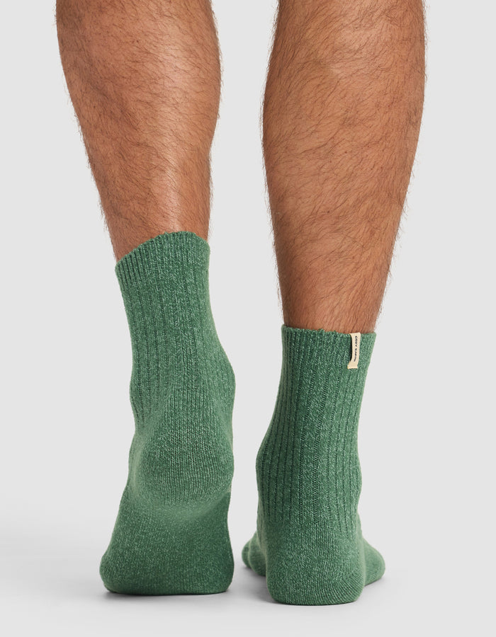 A close-up of someone wearing Cozy Earth’s Plush Lounge Quarter Socks in green, showing ribbed texture and a small fabric tag on the cuff, standing on a flat surface.|Color:Jade/Wintergreen/Pistachio Cream