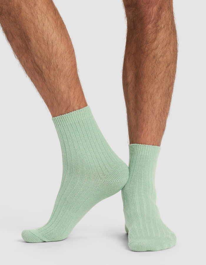 A person with hairy legs is wearing Cozy Earth’s Plush Lounge Quarter Socks in light green, standing with one foot slightly crossed behind the other on a plain white background.|Color:Wintergreen