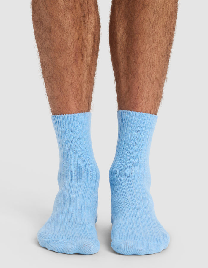 Someone wearing Cozy Earth’s The Plush Lounge Quarter Sock in light blue stands with feet together on a white surface, shown from behind at ankle level.|Color:Harbor