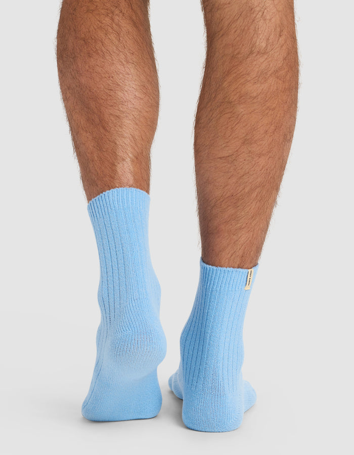 A close-up of a person's lower legs and feet from behind, wearing Cozy Earth’s Plush Lounge Quarter Sock in light blue, set against a plain white background.|Color:Harbor