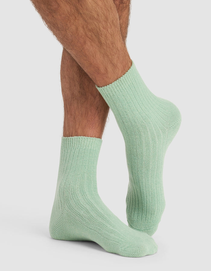 A person with crossed legs is wearing Plush Lounge Quarter Socks by Cozy Earth in light green, shown from the calves down against a plain white background.|Color:Wintergreen